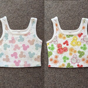 May include: Two white crop tops. One features a pattern of pastel Mickey Mouse silhouettes. The other has a pattern of fruit slices, including watermelon, kiwi, and oranges, in a Mickey Mouse design.