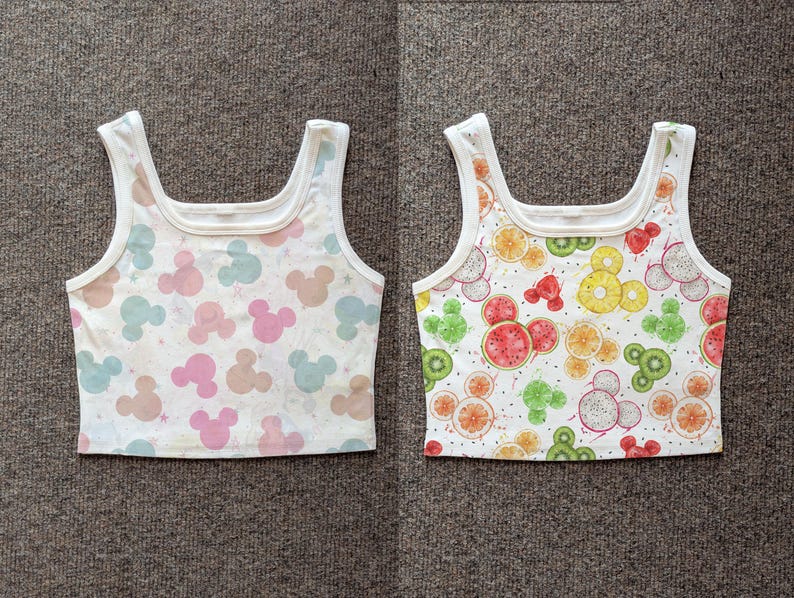 May include: Two white crop tops. One features a pattern of pastel Mickey Mouse silhouettes. The other has a pattern of fruit slices, including watermelon, kiwi, and oranges, in a Mickey Mouse design.