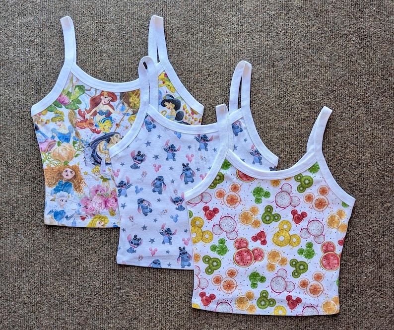 Puede incluir: Tres camisetas sin mangas blancas con tirantes finos. Una camiseta presenta un estampado colorido de princesas de Disney, otra tiene un patr&oacute;n del personaje Stitch, y la tercera muestra un dise&ntilde;o con tem&aacute;tica de frutas con formas de Mickey Mouse.