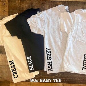 May include: Four 90s baby tee shirts in different colors: cream, black, ash grey, and white. Each shirt has the color name printed on the side.