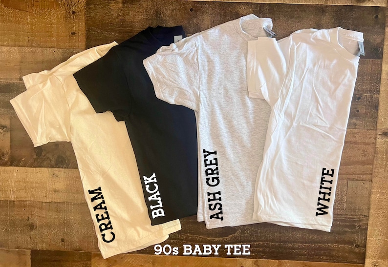 May include: Four 90s baby tee shirts in different colors: cream, black, ash grey, and white. Each shirt has the color name printed on the side.