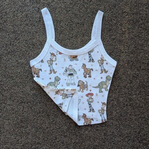 May include: White tank top with a print of characters from the Toy Story movie franchise. The print includes Woody, Buzz Lightyear, Jessie, and other characters.
