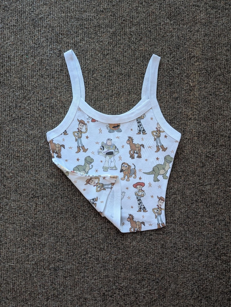 May include: White tank top with a print of characters from the Toy Story movie franchise. The print includes Woody, Buzz Lightyear, Jessie, and other characters.