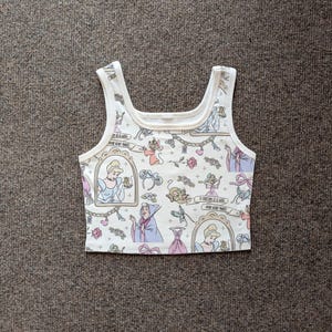May include: White crop top with a repeating pattern of Cinderella-themed illustrations. The design includes Cinderella in a frame, mice, a dress, and text. The top has a scoop neck and wide straps.