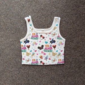 May include: White tank top with a colorful pattern featuring the text "It's a Small World!" and various cartoon icons. The design includes hearts, Mickey Mouse ears, and other whimsical elements. The top has a scoop neckline and wide straps.