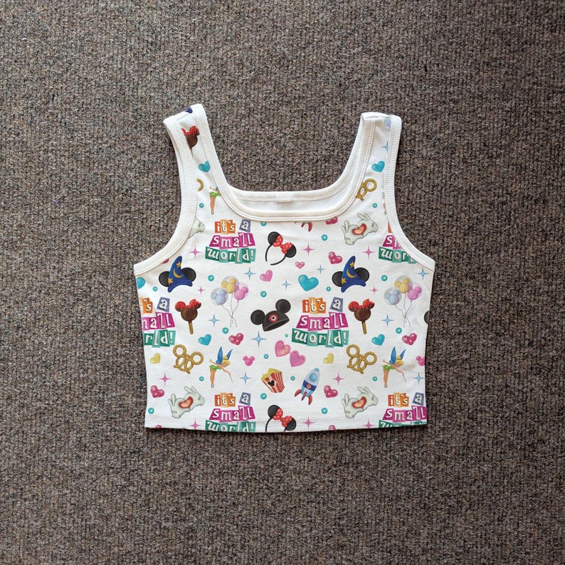 May include: White tank top with a colorful pattern featuring the text "It's a Small World!" and various cartoon icons. The design includes hearts, Mickey Mouse ears, and other whimsical elements. The top has a scoop neckline and wide straps.