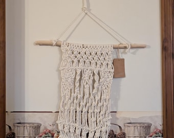 May include: A white macrame wall hanging with a wooden dowel at the top and a brown paper tag hanging from the bottom. The macrame is made with a knot pattern and has long fringe at the bottom.