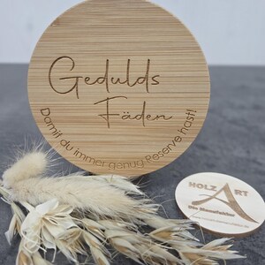 Könnte beinhalten: Ein runder Holzuntersetzer mit den eingravierten Worten "Gedulds Fäden". Der Untersetzer wird von Trockenblumen und einem kleinen Holzschild mit dem Text "HOLZ ART" und einer Website-Adresse begleitet.