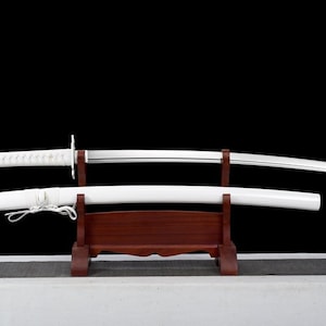 May include: A white katana sword with a white scabbard, displayed on a wooden stand. The sword has a silver blade and a white handle with a chain attached.