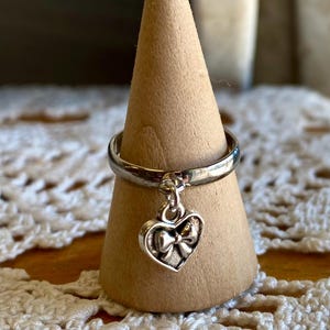 May include: A silver ring with a heart-shaped charm that has a bow on it. The ring is displayed on a wooden ring holder.