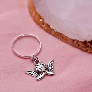 May include: A silver ring with a charm of a cherub with wings hanging from it. The ring is on a pink surface.