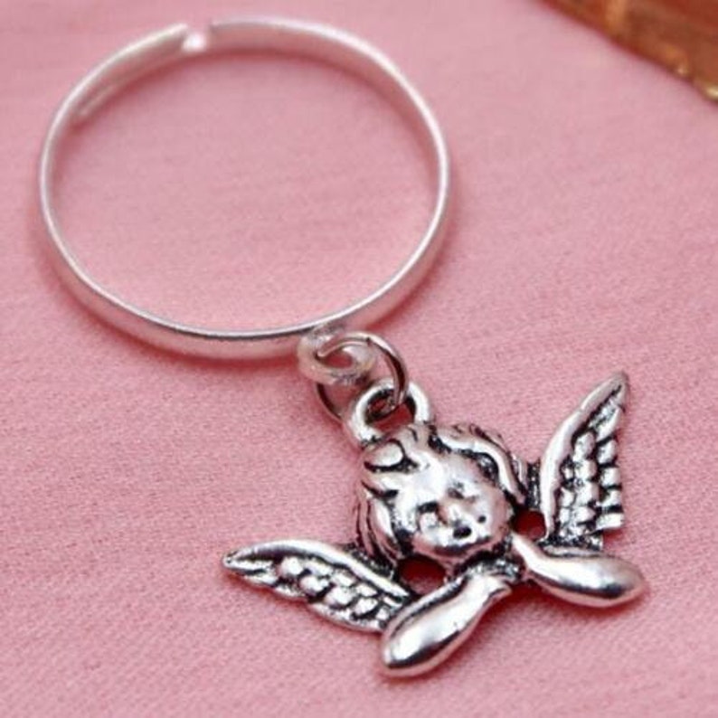 May include: A silver ring with a dangling charm of a silver angel with wings. The angel is looking down with its head resting on its hands.