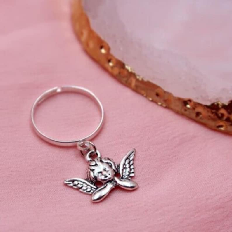 May include: A silver ring with a charm of a cherub with wings hanging from it. The ring is on a pink surface.