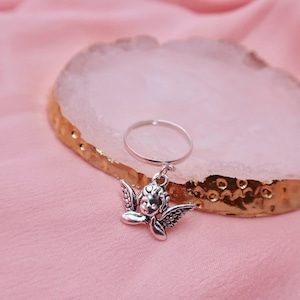 May include: A silver ring with a charm of a cherub angel with wings. The ring is on a pink and gold surface.
