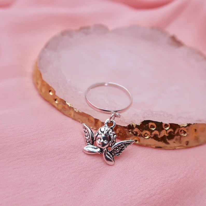May include: A silver ring with a charm of a cherub angel with wings. The ring is on a pink and gold surface.
