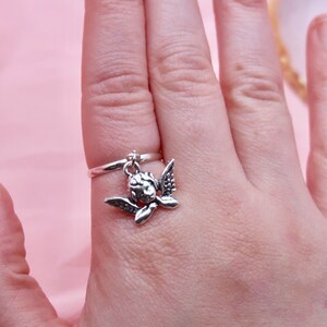 May include: A silver ring with a dangling cherub charm. The cherub is depicted with wings outstretched and a sweet expression.
