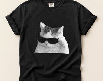 May include: Black t-shirt featuring a halftone image of a cat wearing sunglasses. The cat's image is in black and white, with a star-shaped sparkle on one side. The shirt has rolled-up sleeves and a Comfort Colors tag.
