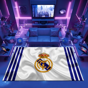Puede incluir: Una alfombra blanca y azul con el escudo del Real Madrid en el centro. El escudo presenta una corona dorada, una letra "M" blanca y una letra "F" blanca dentro de un c&iacute;rculo azul.