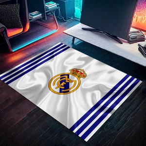 Puede incluir: Alfombra blanca y azul con un logotipo del equipo de f&uacute;tbol del Real Madrid en el centro. El logotipo es un c&iacute;rculo blanco con una corona dorada en la parte superior y las letras "Real Madrid" escritas en oro dentro del c&iacute;rculo.