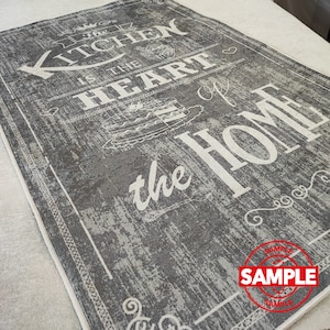 Puede incluir: Una alfombra de cocina gris y blanca con el texto "The Kitchen is the Heart of the Home" en una fuente desgastada. La alfombra tiene un borde blanco y un sello de muestra en la esquina inferior derecha.
