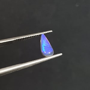 Pode incluir: Uma gema de opala em forma de lágrima com tons azuis e roxos iridescentes, segurada em pinças contra um fundo preto.
