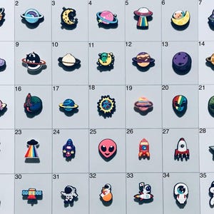 May include: A set of 35 colorful space-themed charms, featuring planets, rockets, astronauts, aliens, and stars. The charms are arranged in a grid pattern on a white background.