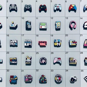 May include: A collection of thirty-five colorful shoe charms arranged in a grid, featuring various gaming-themed designs including video game controllers, handheld consoles, headsets, and text-based slogans like Game Over and Level Up.