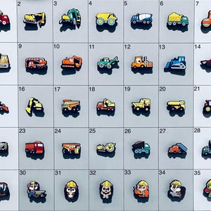 May include: A collection of thirty-five individual shoe charms arranged in a grid, featuring various colorful construction vehicles like dump trucks, excavators, and bulldozers, alongside cartoon characters wearing yellow construction hard hats.