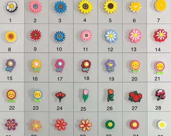 Könnte beinhalten: Fünfunddreißig bunte, blumenförmige Charms mit verschiedenen Designs, darunter Smiley-Gesichter, Herzen und Blumenmuster. Die Charms sind in einem Raster auf weißem Hintergrund angeordnet.