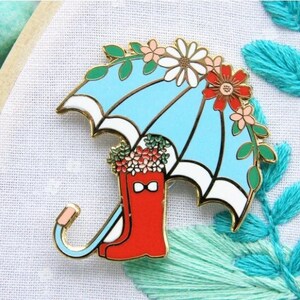 May include: A gold-tone enamel pin featuring a red rain boot with a white bow, filled with colorful flowers, and topped with a blue and white umbrella with floral accents.