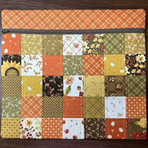 May include: A square zippered pouch featuring a patchwork design of various fabric squares in shades of orange, yellow, brown, and green. The patterns include sunflowers, pumpkins, leaves, and plaid. A solid orange plaid fabric borders the top edge.