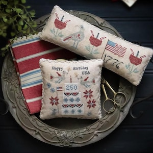 Puede incluir: Una bandeja decorativa contiene varios art&iacute;culos de tem&aacute;tica patri&oacute;tica. Hay dos almohadas bordadas con dise&ntilde;os de p&aacute;jaros y flores, y una bandera americana. Tambi&eacute;n hay una tela a rayas rojas y blancas y unas tijeras doradas. El texto en una almohada dice "Happy Birthday USA 250".