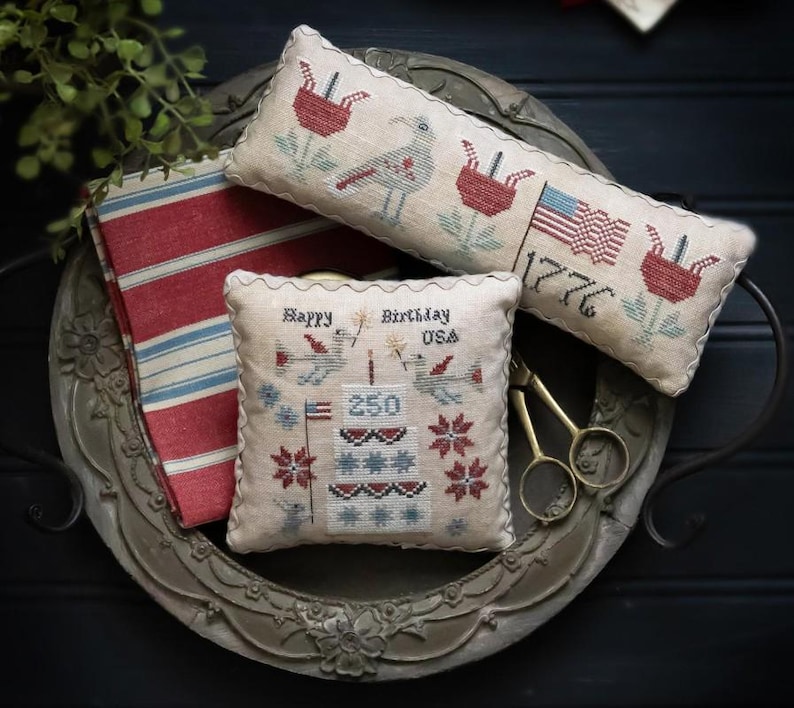 Puede incluir: Una bandeja decorativa contiene varios art&iacute;culos de tem&aacute;tica patri&oacute;tica. Hay dos almohadas bordadas con dise&ntilde;os de p&aacute;jaros y flores, y una bandera americana. Tambi&eacute;n hay una tela a rayas rojas y blancas y unas tijeras doradas. El texto en una almohada dice "Happy Birthday USA 250".