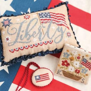 May include: A collection of patriotic cross-stitch items. Includes a pillow with "Liberty" stitched across it, an American flag, and a small floral design. The color scheme is red, white, and blue.
