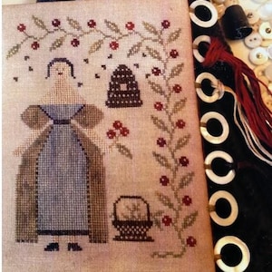 May include: Cross-stitch pattern featuring a woman in a blue dress and brown cloak, standing near a beehive and a basket of flowers. The design is surrounded by a vine with red berries.