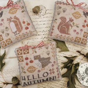 May include: Three decorative, cross-stitched pillows with autumn-themed designs. One pillow features a fox, another a squirrel, and the third reads "Hello Autumn." Each pillow is beige with red and brown accents, tied with a red and white ribbon.