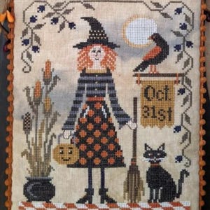 May include: A cross-stitch embroidery on beige fabric featuring a witch with orange hair, a black hat, and a polka-dot skirt. The scene includes a black cat, a crow, a broom, a pumpkin bucket, and a sign reading Oct. 31st, framed by orange pom-poms.