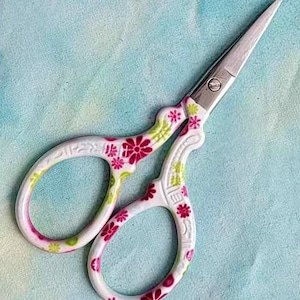 May include: Decorative embroidery scissors with a white handle featuring a floral design in pink and green. The blades are silver and sharp. The scissors are open, ready for use, and rest on a light blue fabric.