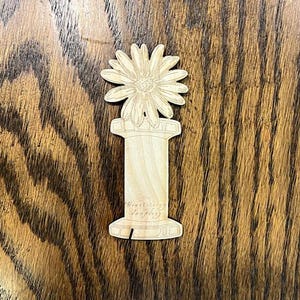 May include: Wooden spool with a flower design on top. The spool is made of light wood and has a simple design.