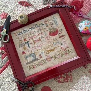 May include: A framed cross-stitch sampler with the text "needles and floss, scissors and spools, these are a stitcher's favourite tools." The red frame surrounds a design with sewing tools. A pair of scissors and a ball of thread are next to the frame.