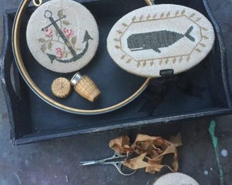 May include: Three embroidery hoops with different designs. The first hoop has a black anchor with pink flowers and green leaves. The second hoop has a black whale with a brown frame. The third hoop has a red heart with a brown and white checkered pattern.
