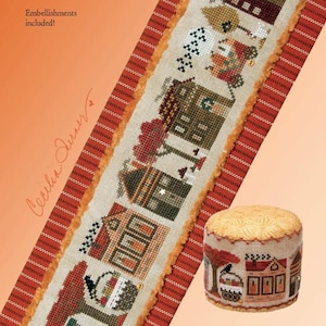 May include: A cross-stitch pattern featuring a harvest-themed tiny town design. The design includes several houses, trees, and other seasonal elements. The image also shows a small, round pincushion with a matching design. The text "Heart in Hand needleart" is visible.