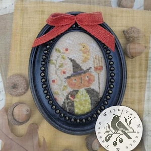 May include: An oval black beaded frame containing a cross-stitch embroidery of a pumpkin-headed figure wearing a witch hat and cape, holding a pitchfork under a crescent moon. A rustic orange burlap bow is attached to the top of the frame.