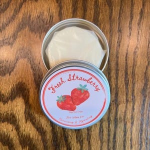 May include: A round, silver tin with a lid featuring a strawberry design and the words "Fresh Strawberry." Inside, a white lotion bar is visible. The tin is open, revealing the product. The background is a wooden surface.