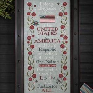 May include: Framed cross-stitch artwork featuring the Pledge of Allegiance. The text is surrounded by floral vines with red flowers. The frame is black with star accents, and the American flag is depicted.