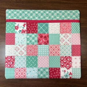 May include: A square fabric pouch featuring a patchwork design of various floral, plaid, and geometric patterns in shades of red, pink, white, and mint green. The top section has a solid mint green border with a bright red zipper closure.