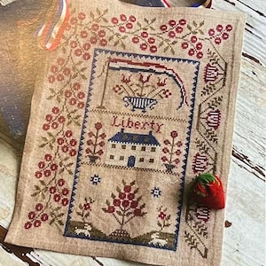 May include: A beige cross-stitch sampler with a patriotic theme, featuring a house, flowers, and the word "Liberty." The design is framed with a floral border and includes a red, white, and blue ribbon. A red strawberry pincushion rests on the corner.