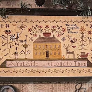 May include: A framed cross-stitch sampler with a festive theme. The design features a yellow house, deer, and floral elements, with the text "A Yuletide Welcome to Thee." The sampler is framed in a dark wooden frame.