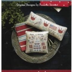 Puede incluir: Una exhibici&oacute;n de tres cojines decorativos con temas patri&oacute;ticos. Un coj&iacute;n presenta un dise&ntilde;o de la bandera estadounidense, otro tiene dise&ntilde;os de flores rojas, y el tercero muestra el texto "Happy Birthday, USA" y el n&uacute;mero "250". Los cojines est&aacute;n sobre una bandeja decorativa.