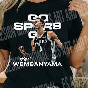 Könnte beinhalten: Ein schwarzes T-Shirt mit einer Grafik des Basketballspielers Victor Wembanyama in einem schwarz-weißen Spurs-Trikot. Der Text GO SPURS GO ist in Weiß über dem Spieler gedruckt, und der Name WEMBANYAMA ist in Weiß am unteren Rand gedruckt.